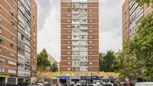A multi-story residential building with balconies, a ground-level commercial area with shops, and parked cars.