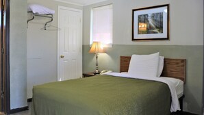 Cama bem arrumada com colcha verde, luminária de cabeceira, quadro na parede e porta branca com toalha pendurada na maçaneta.