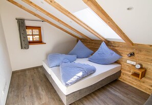 Un dormitorio tipo loft con techo inclinado, vigas de madera, tragaluces, cama con ropa de cama a cuadros, una mesita de noche con un reloj y un pequeño estante.