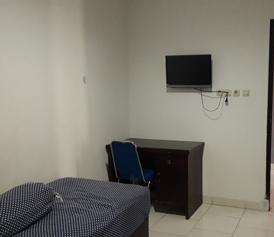 A small, sparsely furnished room with a bed, a desk, and a chair. A television is mounted on the wall above the desk.