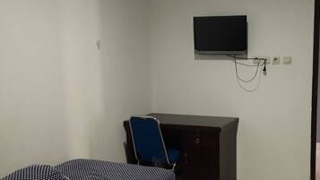 A small, sparsely furnished room with a bed, a desk, and a chair. A television is mounted on the wall above the desk.