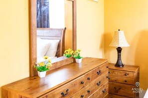 Une commode en bois avec de nombreux tiroirs et un miroir au-dessus. On y voit deux petits pots blancs ornés de fleurs jaunes. Une lampe avec un abat-jour à motifs est posée sur une table de chevet en bois, à côté de la commode.