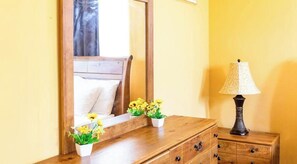 A wooden dresser with multiple drawers and a mirror above it. There are two small white pots with yellow flowers on the dresser. A lamp with a patterned lampshade is on a wooden nightstand next to the dresser.