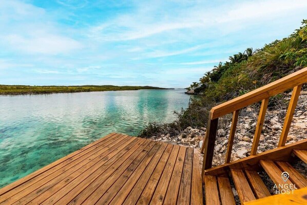 Une terrasse en bois mène à une étendue d’eau calme aux eaux turquoise et limpides, entourée de verdure et sous un ciel dégagé.