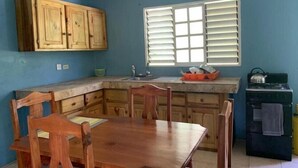A kitchen with wooden cabinets, a dining table, chairs, a window with blinds, and a stove. - Tha Lagoon Spot (Port Antonio)