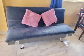 Un sofá azul oscuro con dos cojines rosados, una silla de madera y un suelo de baldosas.
