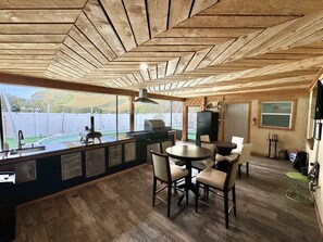 A kitchen with a large island, a dining area with chairs, a TV mounted on the wall, and a view of a backyard with a pool.