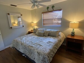 Une chambre à coucher comprenant un lit, deux tables de chevet, un ventilateur de plafond, une fenêtre avec des stores, un miroir et une lampe fixée au mur.