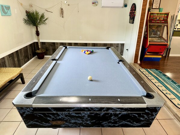 A pool table with balls, a potted plant, and a claw machine in a room with wooden flooring and a tiled wall.