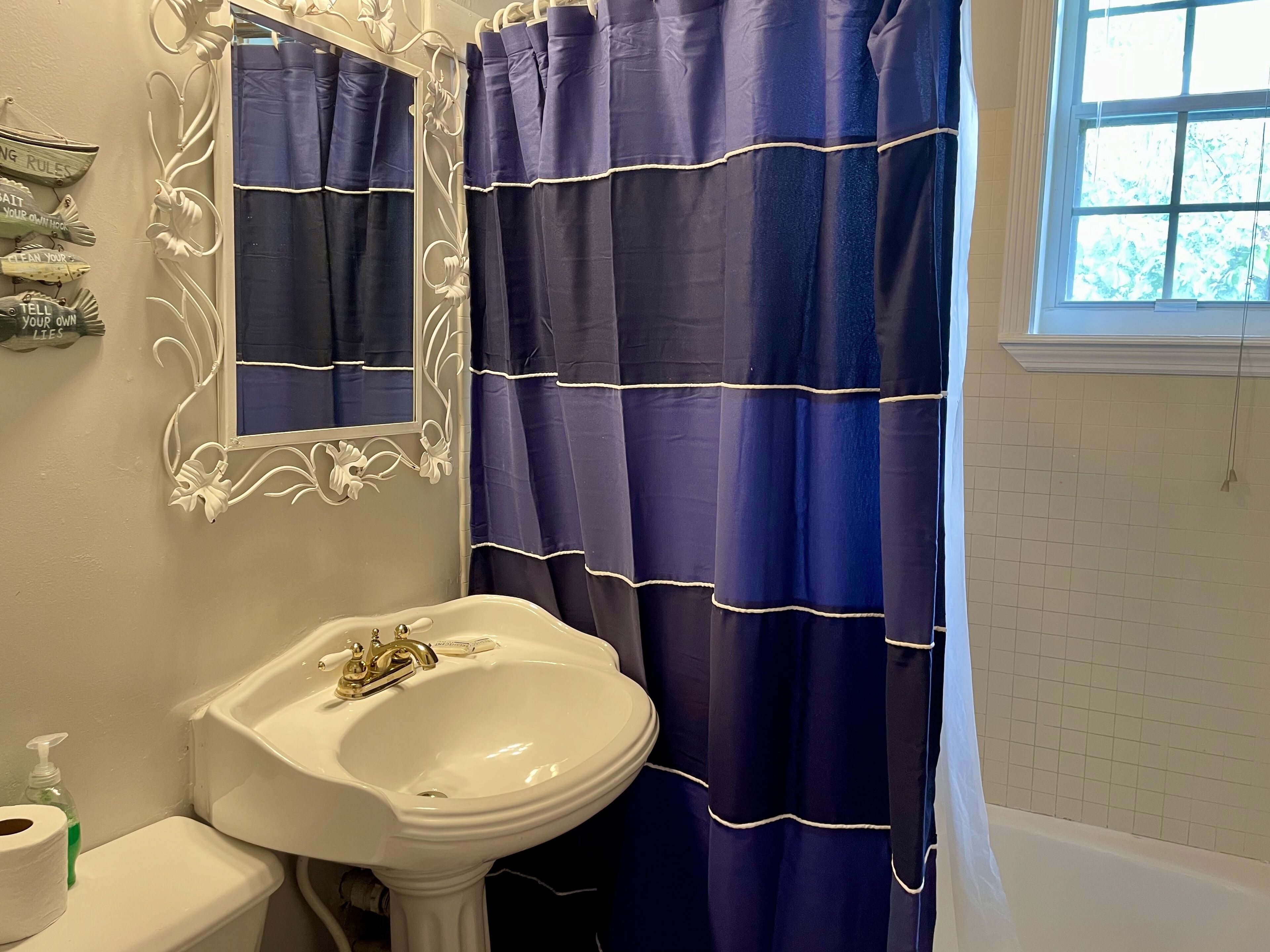 A bathroom with a white pedestal sink, a mirror with decorative molding, a blue shower curtain, and a window with a view of trees.