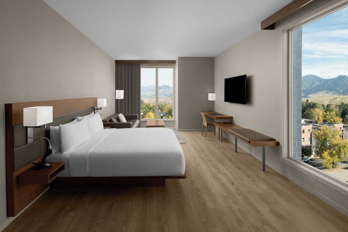 A modern hotel room with a large bed, a desk, a TV, and a view of mountains.