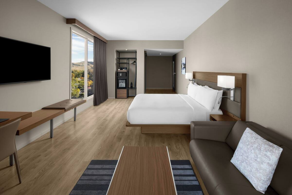 A modern hotel room with a large bed, a desk, a TV, a sofa, and a view of the outdoors.