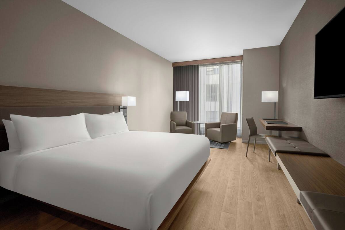 A hotel room with a large bed, two armchairs, a small table, a desk, and a TV mounted on the wall.