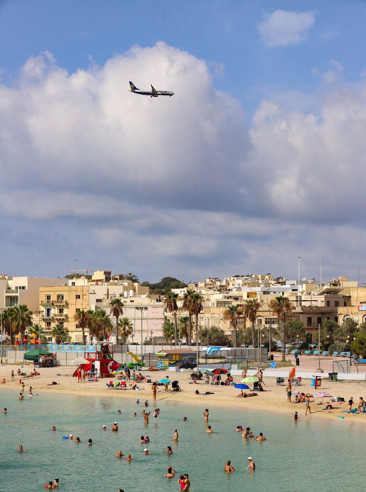 Ein Strand mit badenden und sonnenden Menschen, ein Flugzeug am Himmel und Gebäude im Hintergrund.