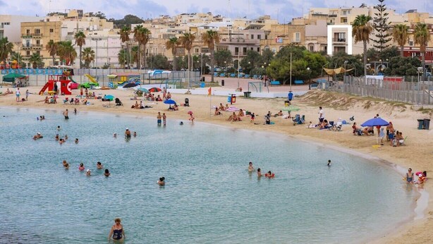 Una playa con gente nadando y tomando el sol, una torre de salvavidas y edificios al fondo.