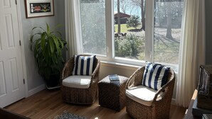 Ein Zimmer mit zwei Korbstühlen mit gestreiften Kissen, einer Topfpflanze, einem gemusterten Teppich und Blick auf ein Haus durch große Fenster.