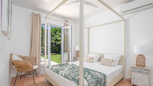 A four-poster bed with a green leaf-patterned bedspread, a wicker chair with cushions, a small bedside table with a lamp, and a view of a garden through a sliding glass door. - Colibri Trotteur (Le Diamant)