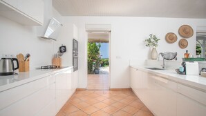 A modern kitchen with white cabinets, a tiled floor, and a view of a garden through an open doorway. - Colibri Trotteur (Le Diamant)
