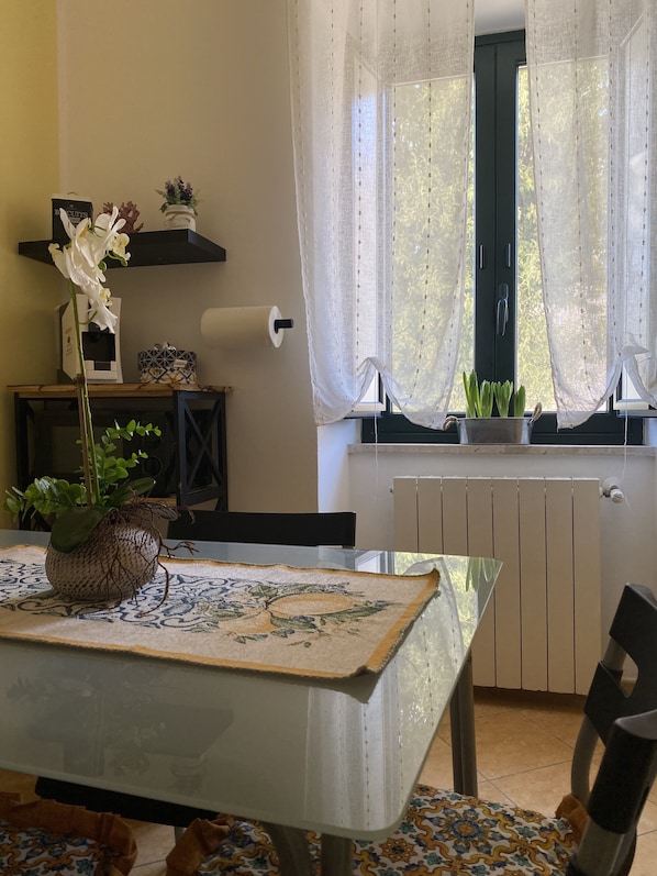 A dining area with a table set for two, a potted plant, and a window with sheer curtains.