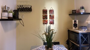 Eine Küche mit einer Kaffeemaschine, einem Wasserkocher und einem Regal mit einem 'Home'-Schild. An der Wand befinden sich Bilder und ein kleiner Topfpflanze.