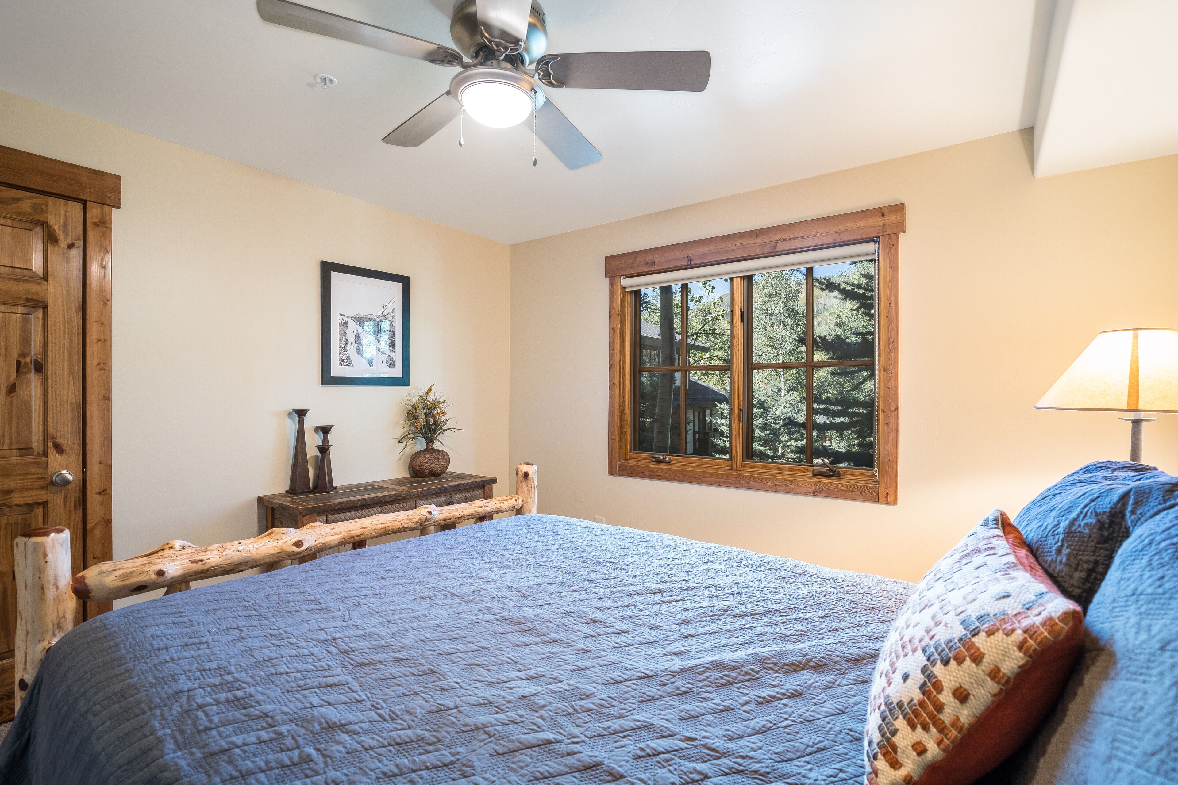 A bedroom with a bed, a ceiling fan, a window with a view of trees, and a wooden headboard.