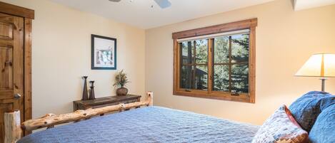 A bedroom with a bed, a ceiling fan, a window with a view of trees, and a wooden headboard.