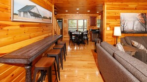 A wooden cabin interior with a long dining table, bar stools, a sofa, and a kitchen area.