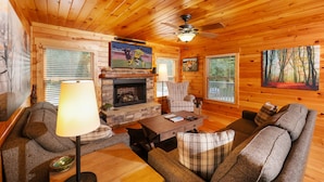 A cozy cabin interior with a fireplace, wooden walls, a ceiling fan, and a television mounted above it.