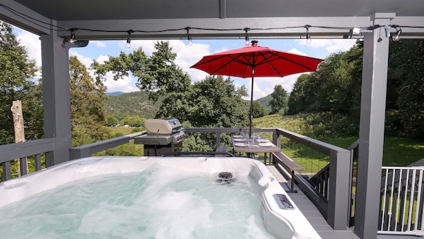 A hot tub on a deck with a red umbrella, surrounded by greenery and a view of hills.