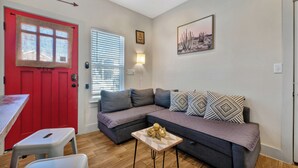 Un espacio habitable pequeño con una puerta roja, un sofá gris, una mesita con un objeto decorativo y una ventana con persianas.