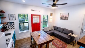 Un espacio habitable pequeño con kitchenette, una puerta roja, un sofá gris y un ventilador de techo.