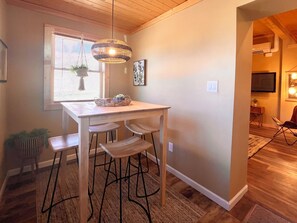 A dining area with a wooden table and bar stools, a hanging light fixture, and a window with blinds.