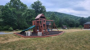 A playground with a slide and climbing structures, surrounded by a grassy area and a wooden fence. - Turkey Roost At Creekside Resort (Clearville)