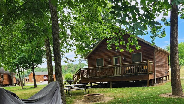 A rustic cabin with a deck, surrounded by trees and a grassy area with firewood. - Turkey Roost At Creekside Resort (Clearville)