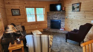 Een gezellig interieur van een blokhut met een stenen openhaard, houten wanden, een televisie erboven en een comfortabele zithoek met een gestreept fauteuil en een bank.