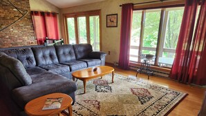 Ein Wohnzimmer mit einer Ledersofa, einem Holztisch und einem gemusterten Teppich. Große Fenster mit Vorhängen, eine Ziegelwand und ein gerahmtes Bild sind ebenfalls zu sehen.
