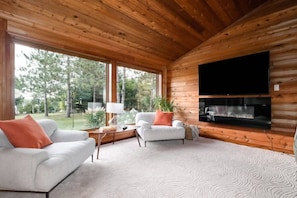 A modern log cabin interior with a fireplace, two white sofas, a flat-screen TV, and large windows offering a view of the surrounding forest. - Xanadu - Zen waterfront retreat on Upper Rideau (Westport)