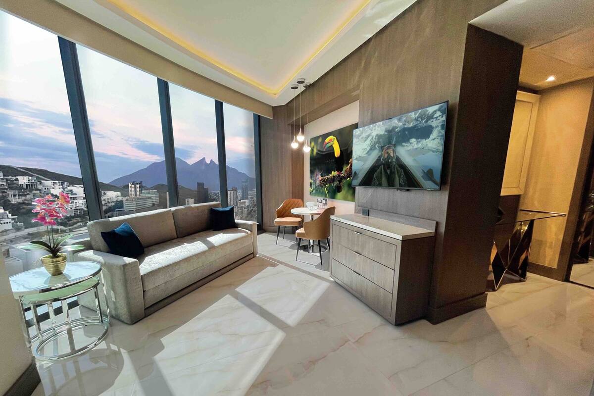 Een moderne woonkamer met een bank, salontafel en eethoek. Een grote televisie is aan de muur bevestigd, en er is uitzicht op de stad door grote ramen.