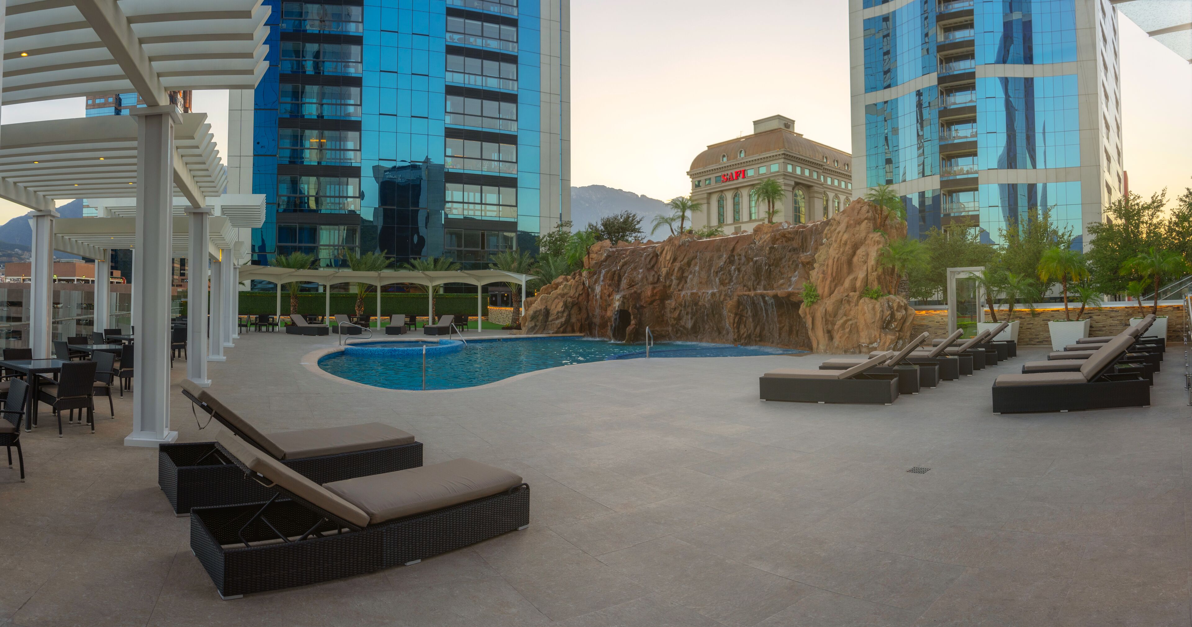 Un área de piscina en la azotea con sillones de descanso, una cascada y edificios modernos al fondo.