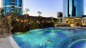 A swimming pool with a rock formation and palm trees, surrounded by modern buildings. - Safi Royal Luxury Metropolitan (San Pedro Garza García)