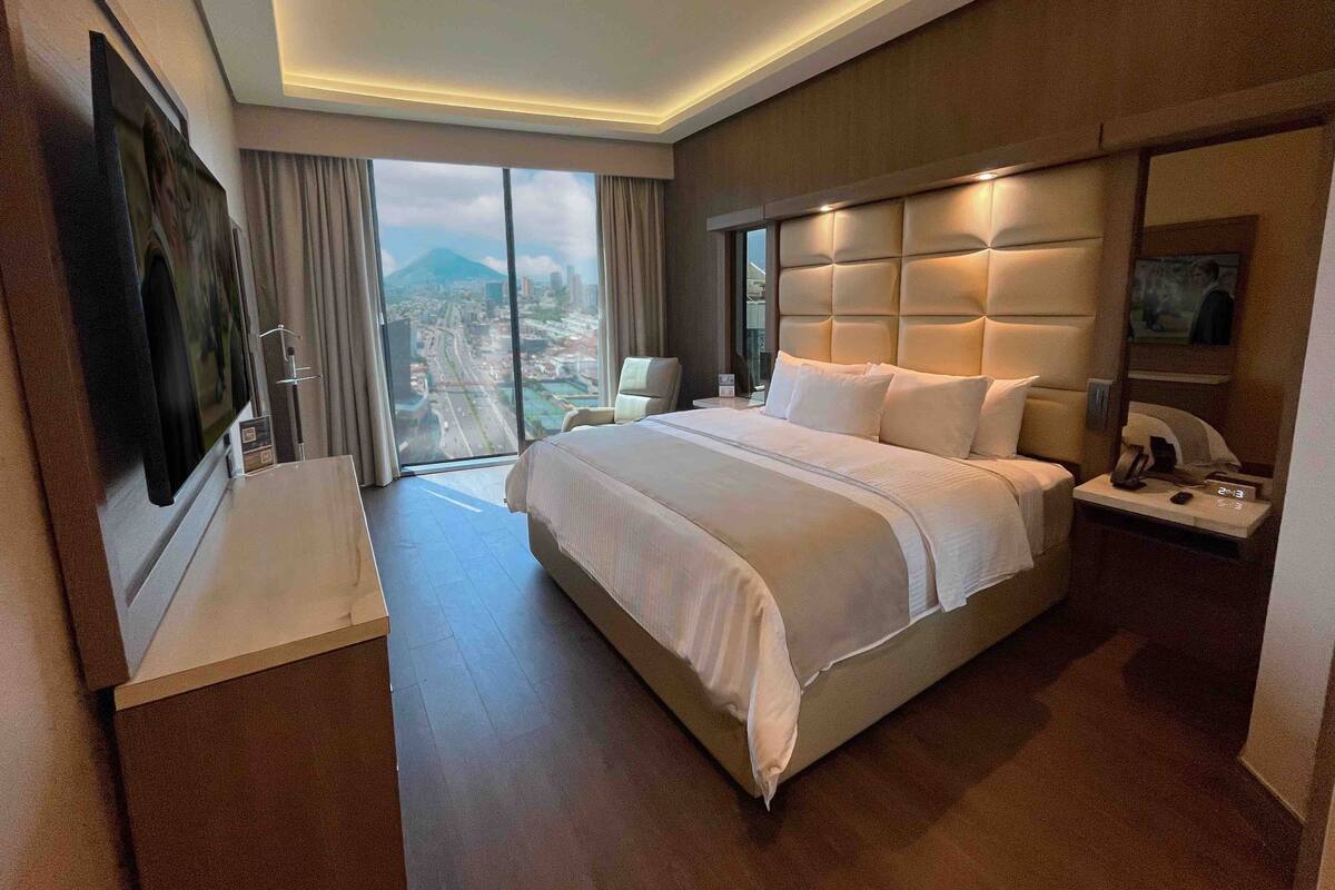 Een moderne hotelkamer met een groot bed, een flatscreen-tv, een bureau met telefoon en uitzicht op de stad door grote ramen.