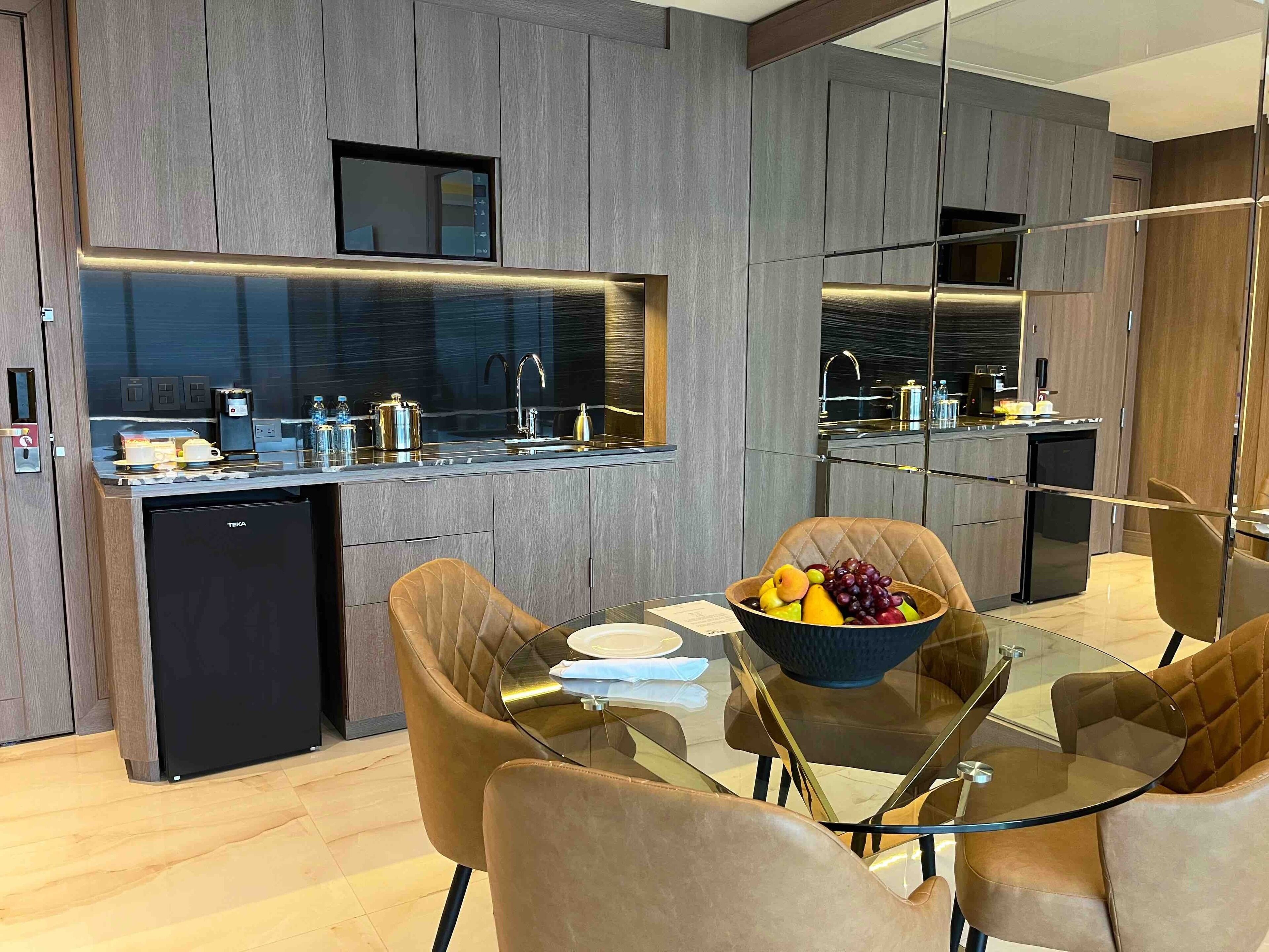 A modern kitchen with a built-in oven, a coffee machine, and a sink. There is a dining area with a glass table and leather chairs.