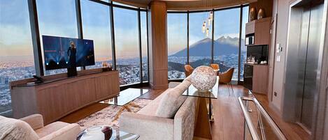 Una sala de estar moderna con un televisor grande, asientos cómodos y vistas a un paisaje urbano y montañas.
