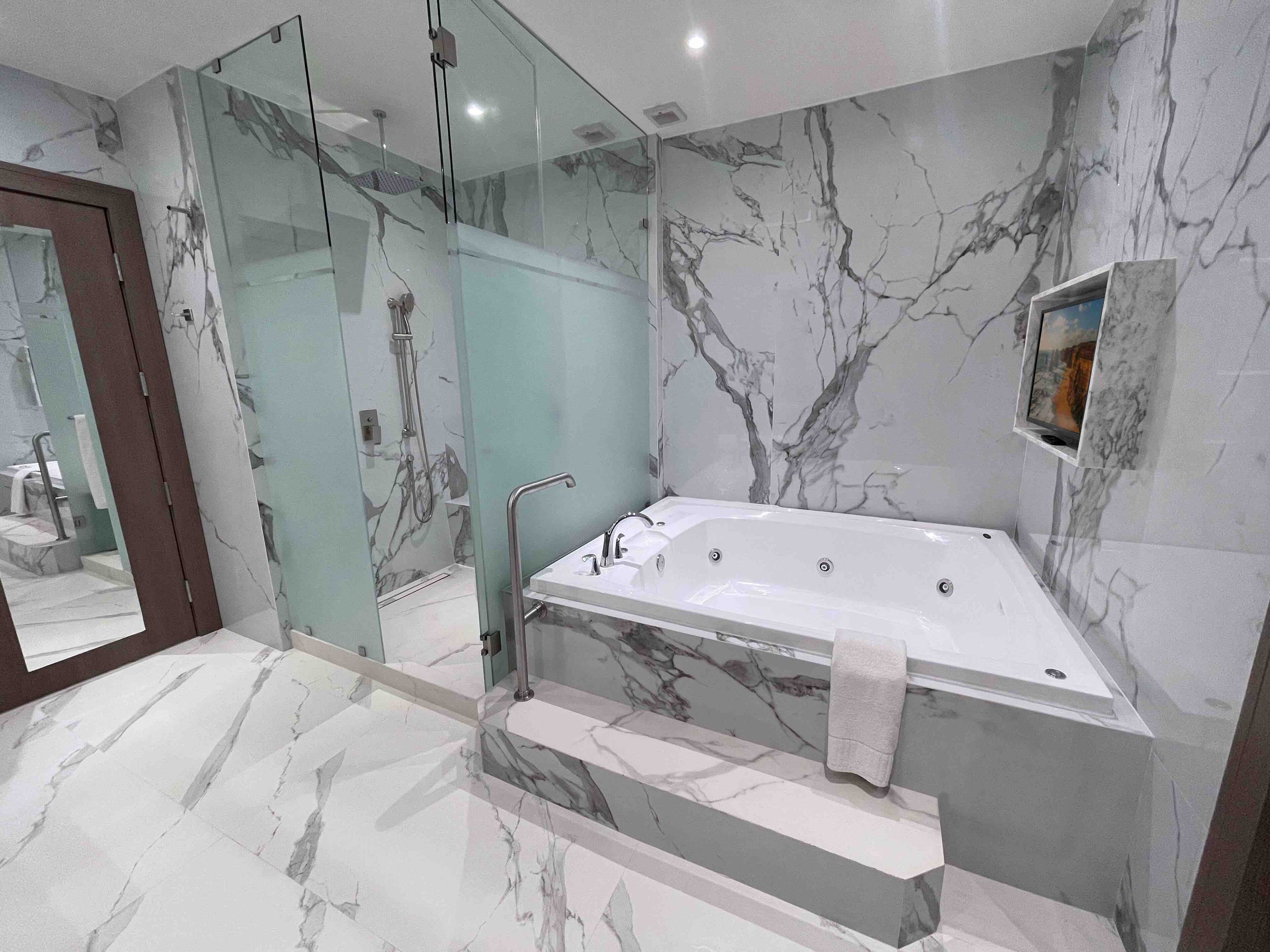 Un baño moderno con suelo de mármol, ducha con mampara de vidrio, bañera independiente y un pequeño televisor montado en la pared.