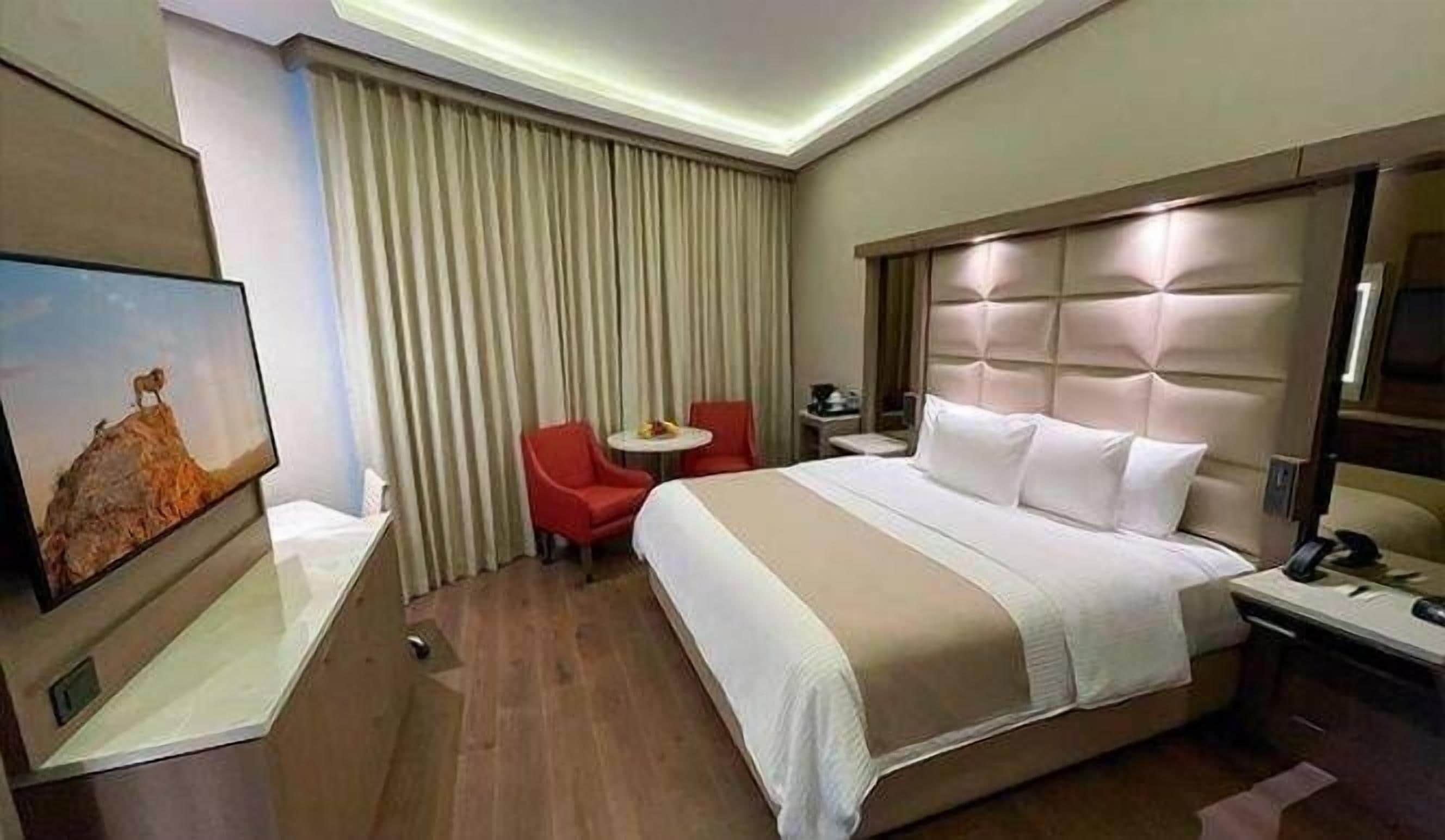 A hotel room with a large bed, a small table with chairs, a TV, and a window with curtains.