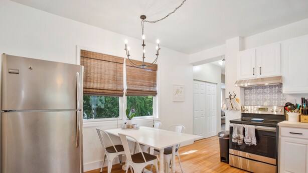 A modern kitchen with a stainless steel refrigerator, white cabinetry, a wooden dining table with chairs, and a large window with blinds.