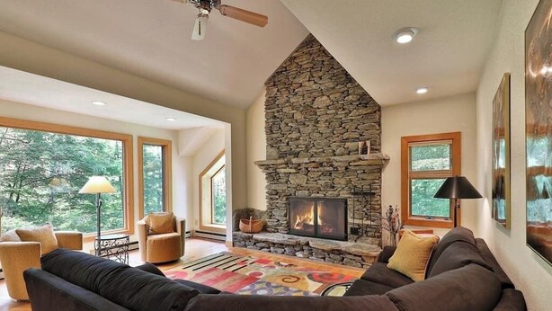 A living room with a stone fireplace, large windows, and a ceiling fan.