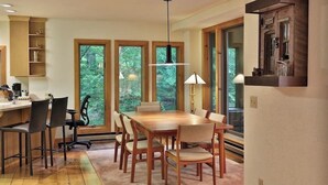 A dining area with a wooden table and chairs, large windows, and a hanging light fixture.