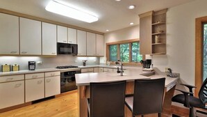 A modern kitchen with white cabinets, a central island with chairs, and a window overlooking greenery.