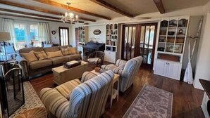 A cozy living room with a sofa, armchairs, a coffee table, and a chandelier. There are bookshelves and a television in the background. - Twin River Stone Cottage - 7 BR/Sleeps 20 (Norfork)